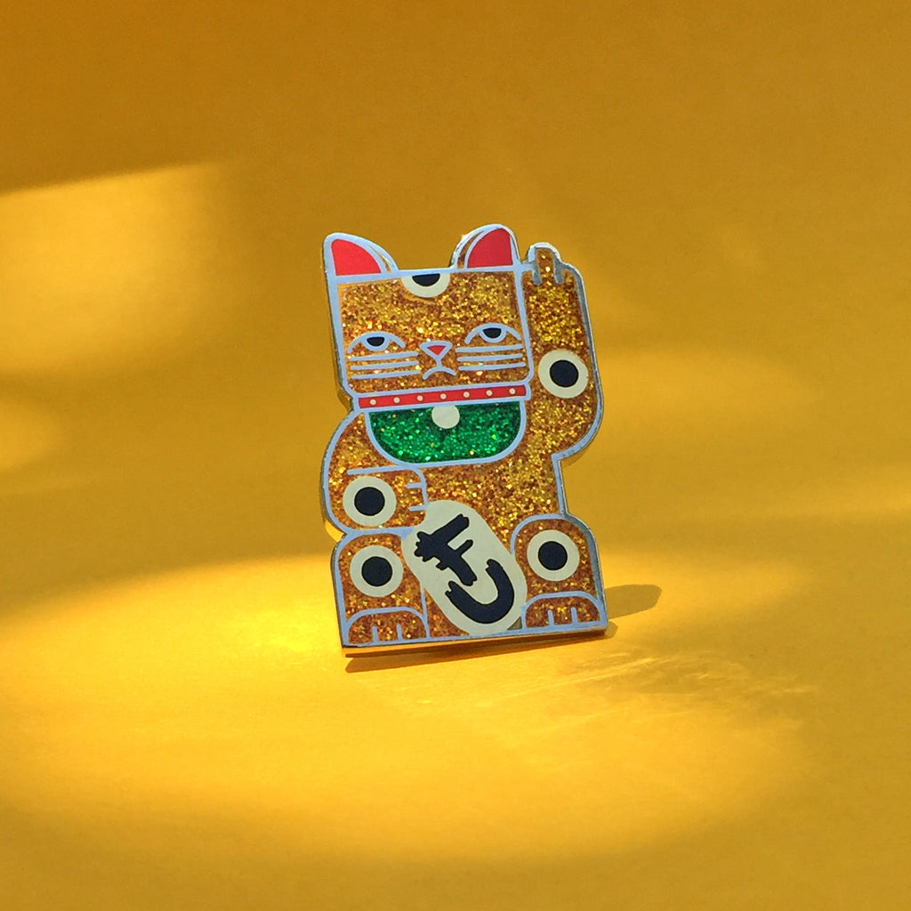 Goodbye Kitty gold glitter enamel pin in gold and silver with green glitter bib. Shows a maneki neko rudely gesturing.