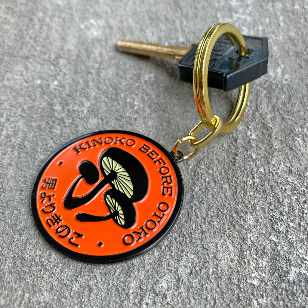 Kinoko Before Otoko, mushrooms before dudes enamel split-ring keychain in orange and black with key.
