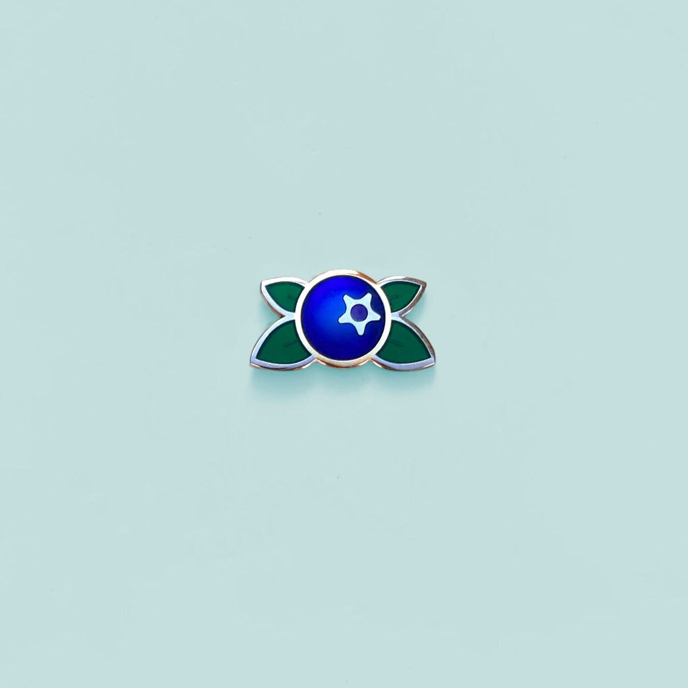 Small blue and green blueberry enamel pin on a light blue background