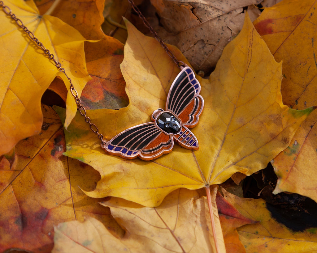 Atropos enamel death's-head moth necklace in copper seconds with black mirrored siofourite crystal skull among autumn leaves.