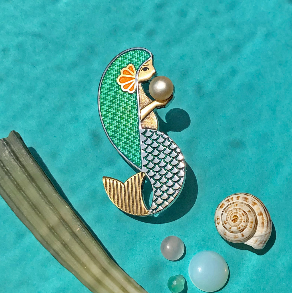 Amathea enamel mermaid pin in aqua with freshwater pearl laying among seashells.