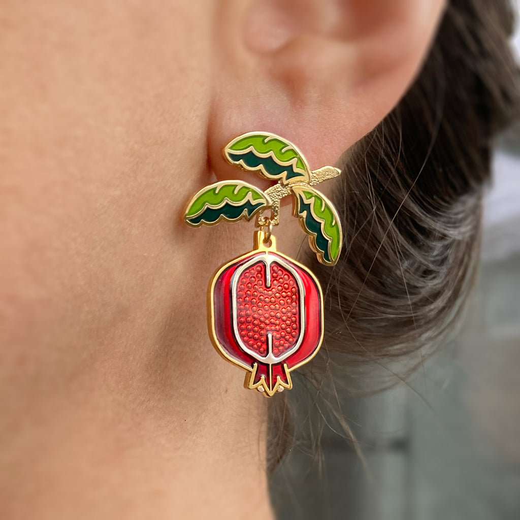 A person wearing a Pomegranate enamel earring, shows the fruit dangle below the earlobe under the leaf branch stud.