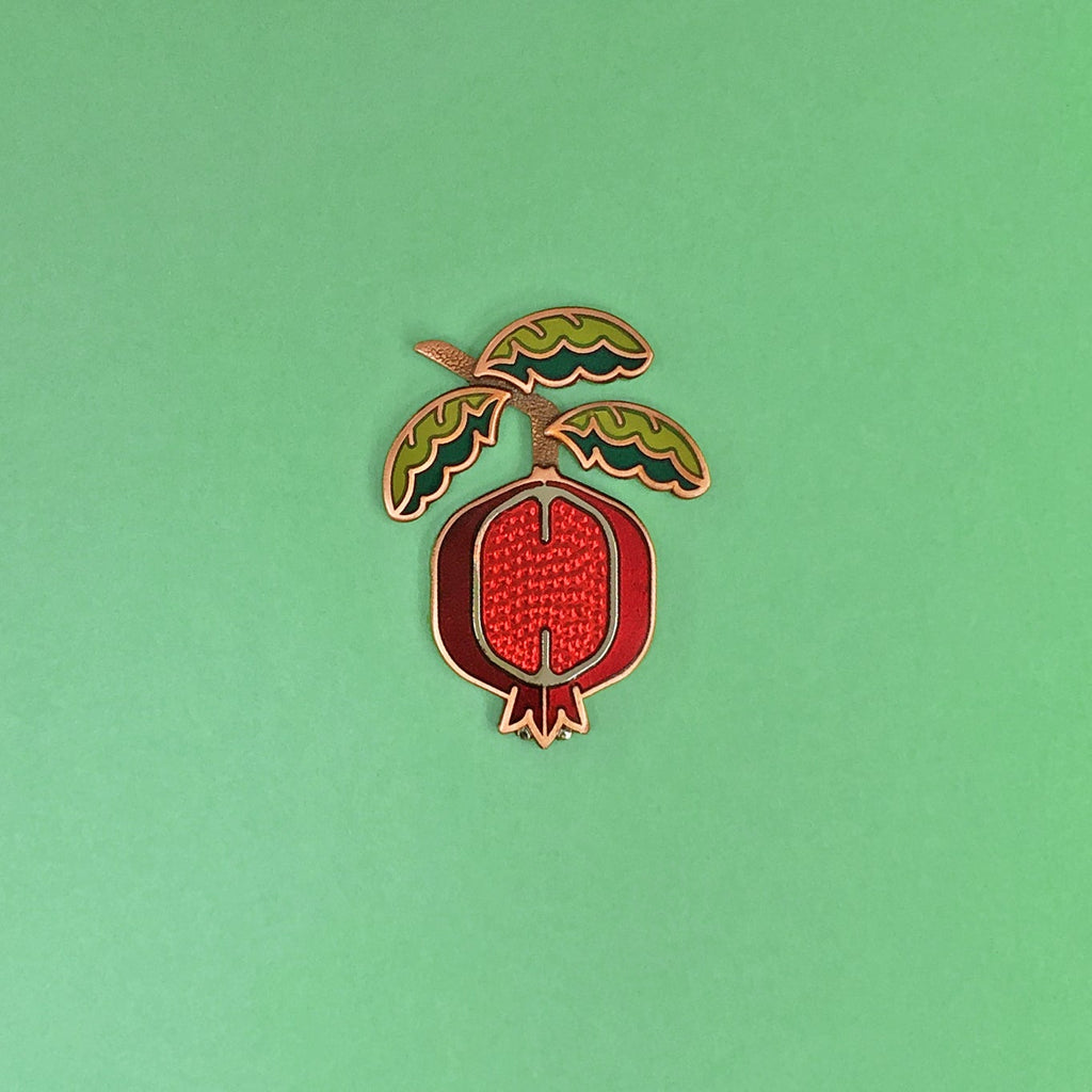 Pomegranate enamel pin seconds in silver and antique copper with green enamel leaf branch and red sliced fruit.