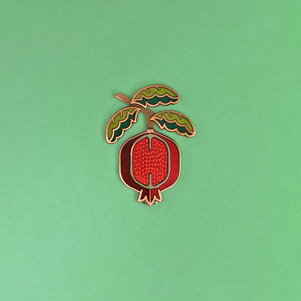 Pomegranate enamel pin in silver and antique copper with green enamel leaf branch and red sliced fruit.