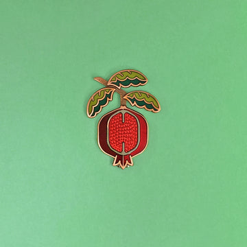 Pomegranate enamel pin in silver and antique copper with green enamel leaf branch and red sliced fruit.