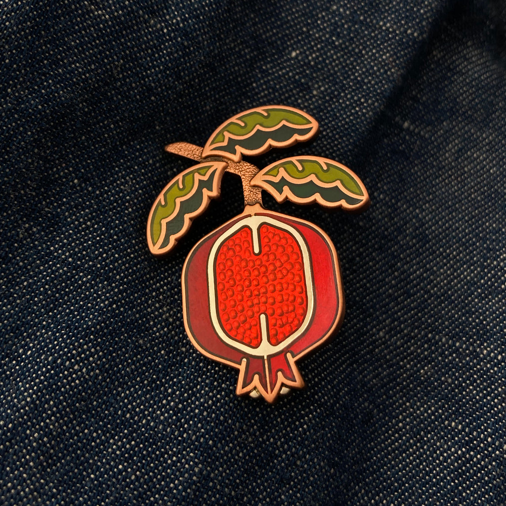 Pomegranate pin in silver and antique copper on a jean jacket with green enamel leaf branch and red sliced fruit.