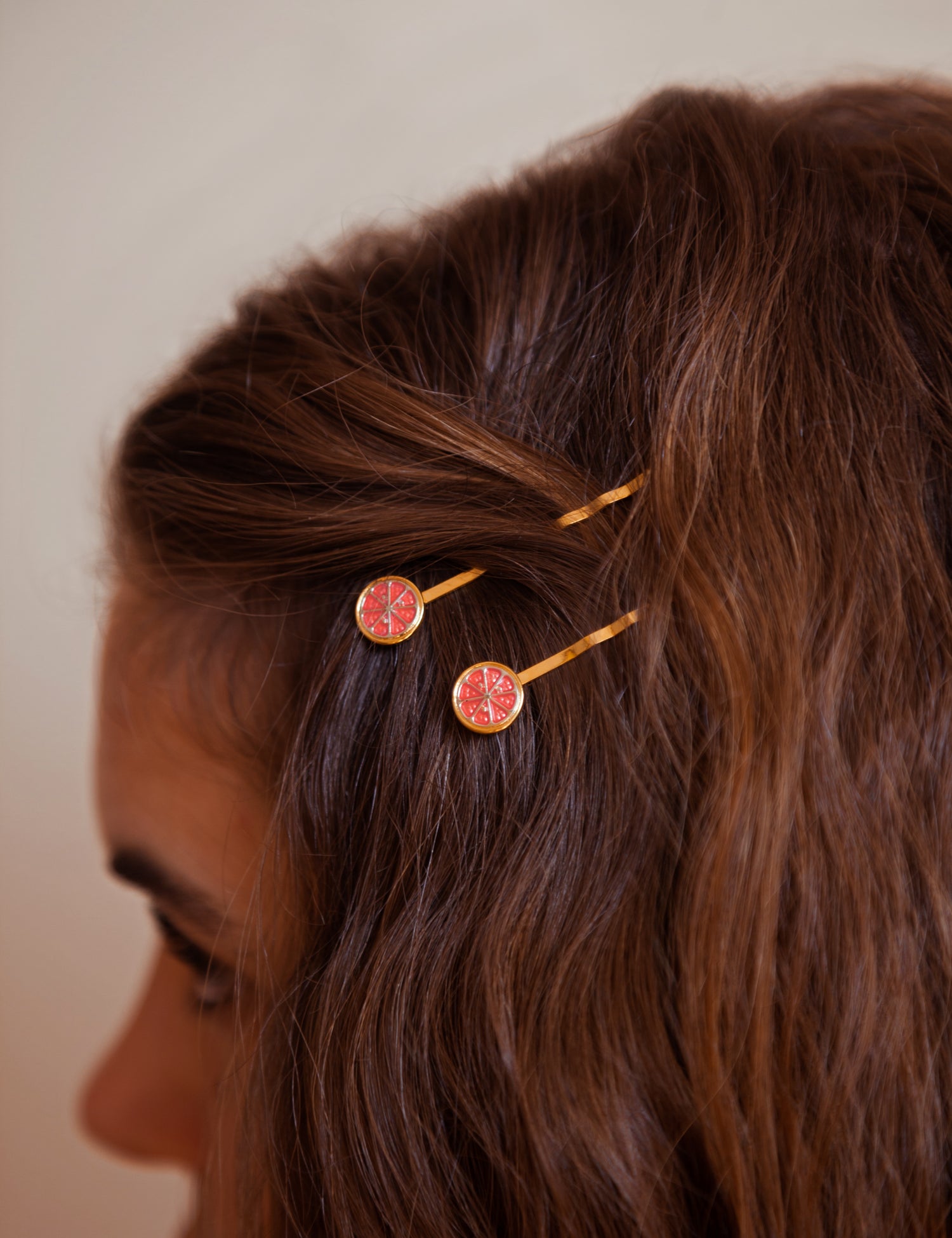 A person wearing two Ruby Red grapefruit hairpins finished in 24k gold and sterling silver with red enamel sliced fruit.