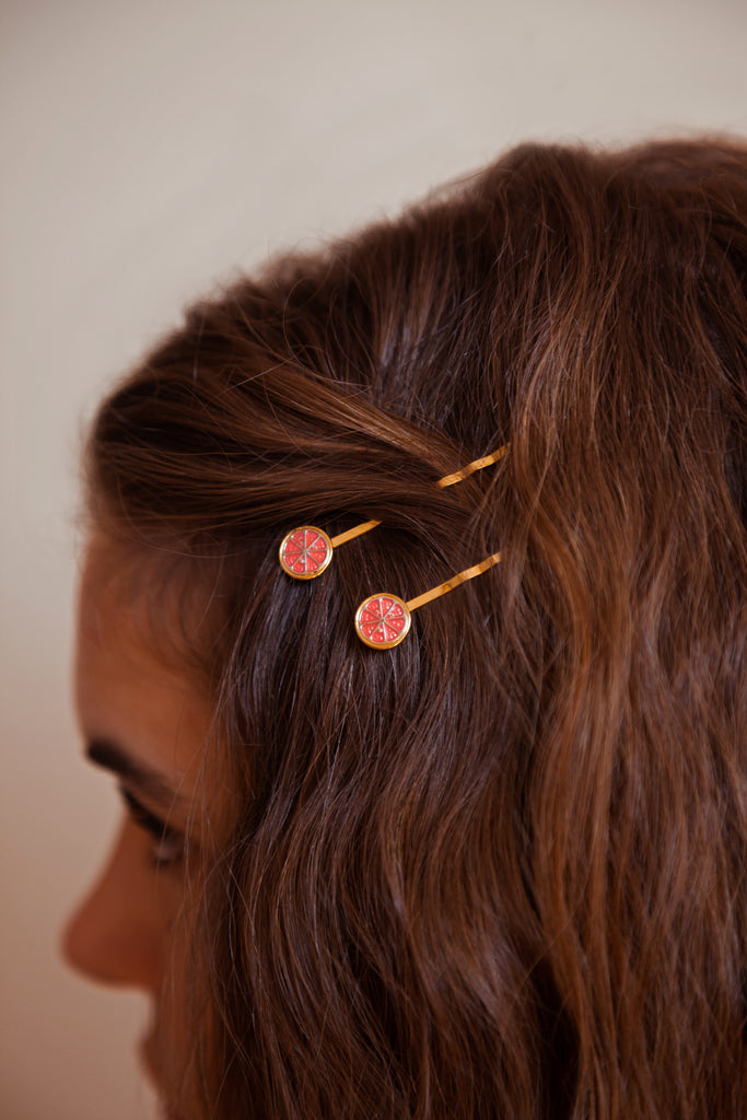 A person wearing two Ruby Red grapefruit hairpins finished in 24k gold and sterling silver with red enamel sliced fruit.