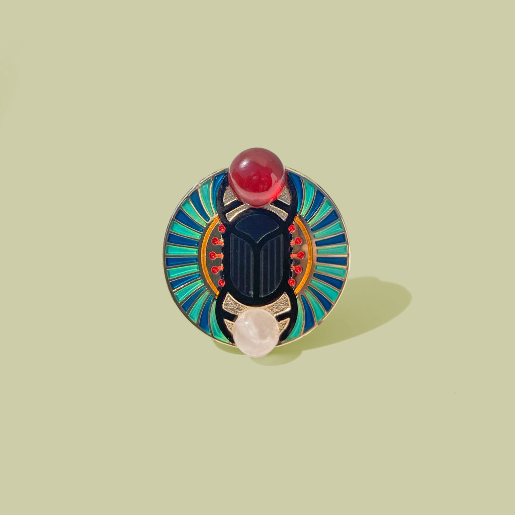 Scarab enamel pin with gold and black finish, blue sunburst border and carnelian and rose quartz cabochons.